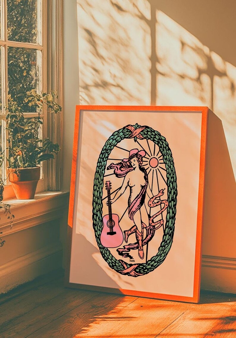 K&ouml;nnte beinhalten: Eine gerahmte Illustration einer Frau mit Cowboyhut, die eine rosafarbene Gitarre h&auml;lt. Sie ist von einem gr&uuml;nen Kranz und einer Schlange umgeben. Der Hintergrund ist pfirsichfarben.
