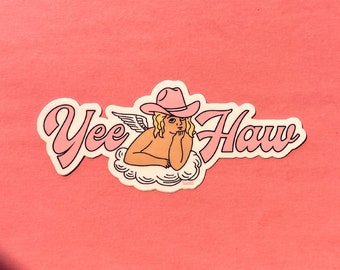 Può includere: Un adesivo con un angelo che indossa un cappello da cowboy rosa con le ali, che riposa su una nuvola bianca, con il testo "Yee Haw" scritto in grassetto.