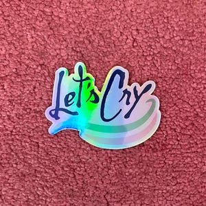 Peut inclure: Un autocollant holographique avec le texte "Let's Cry" en &eacute;criture noire. L'autocollant a une finition arc-en-ciel iris&eacute;e.