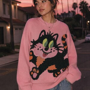 May include: A long-sleeve pink knit sweater featuring a large graphic of a black cat with green eyes, a pink face, and orange accents on its paws and tail. The sweater has a crew neckline and a relaxed fit, paired with blue denim jeans.
