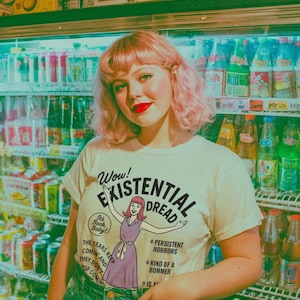 May include: A woman wearing a cream-colored t-shirt with the words "Wow! Existential Dread" and a cartoon illustration. She is standing in front of a refrigerator filled with beverages. She is wearing blue jeans and has pink hair.