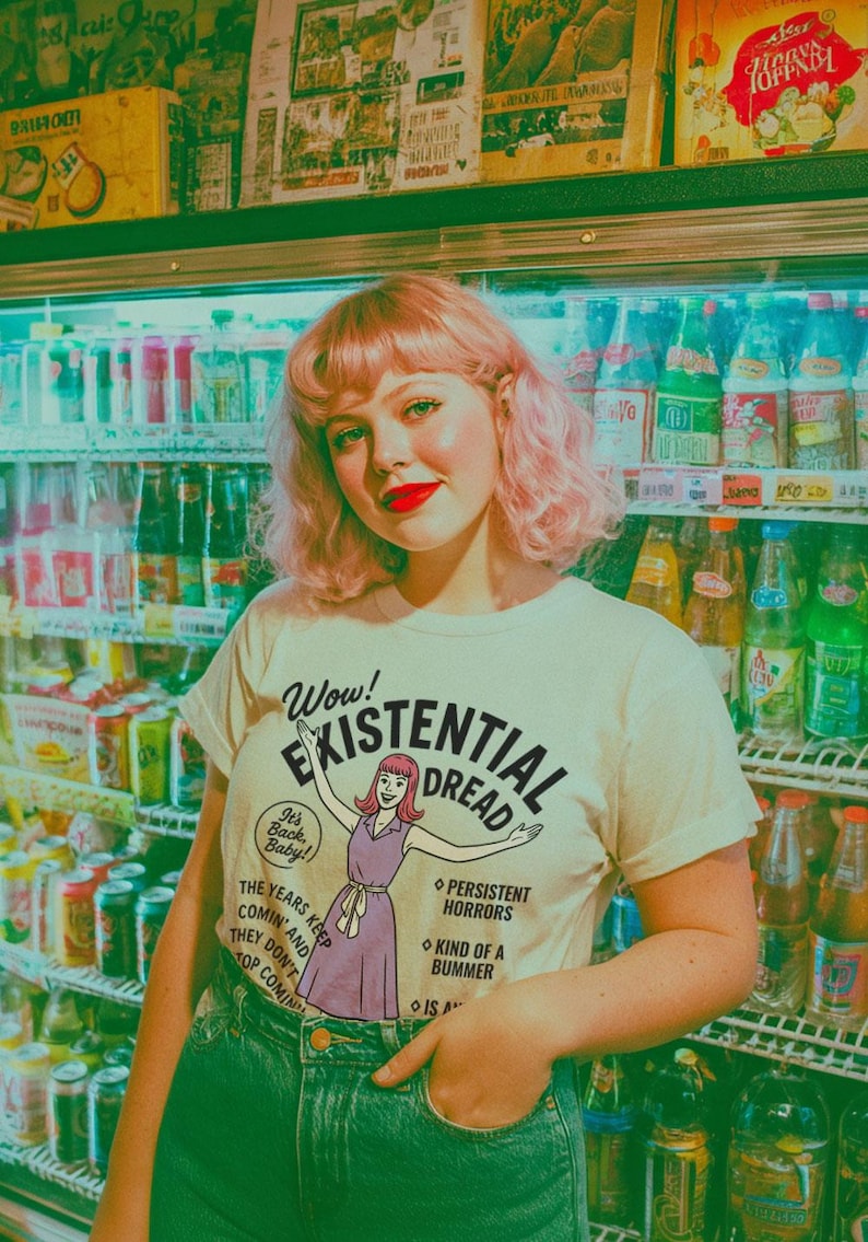 May include: A woman wearing a cream-colored t-shirt with the words "Wow! Existential Dread" and a cartoon illustration. She is standing in front of a refrigerator filled with beverages. She is wearing blue jeans and has pink hair.