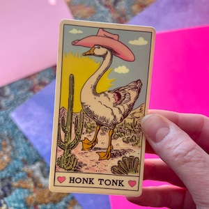 Könnte beinhalten: Eine skurrile Karte mit einer Gans, die einen rosa Cowboyhut trägt, vor einer Wüstenkulisse mit Kakteen. Auf der Karte steht der Text "HONK TONK", mit einem Herzsymbol auf beiden Seiten des Satzes.
