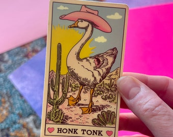 Peut inclure: Carte fantaisie avec une oie portant un chapeau de cow-boy rose, dans un décor désertique avec des cactus. Le texte "HONK TONK" est imprimé sur la carte, avec un cœur de chaque côté.