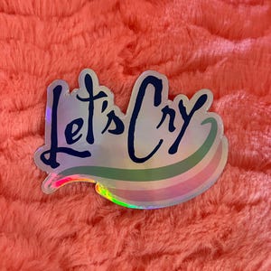 Peut inclure: Un autocollant holographique avec un motif arc-en-ciel et le texte "Let's Cry" en lettres noires.