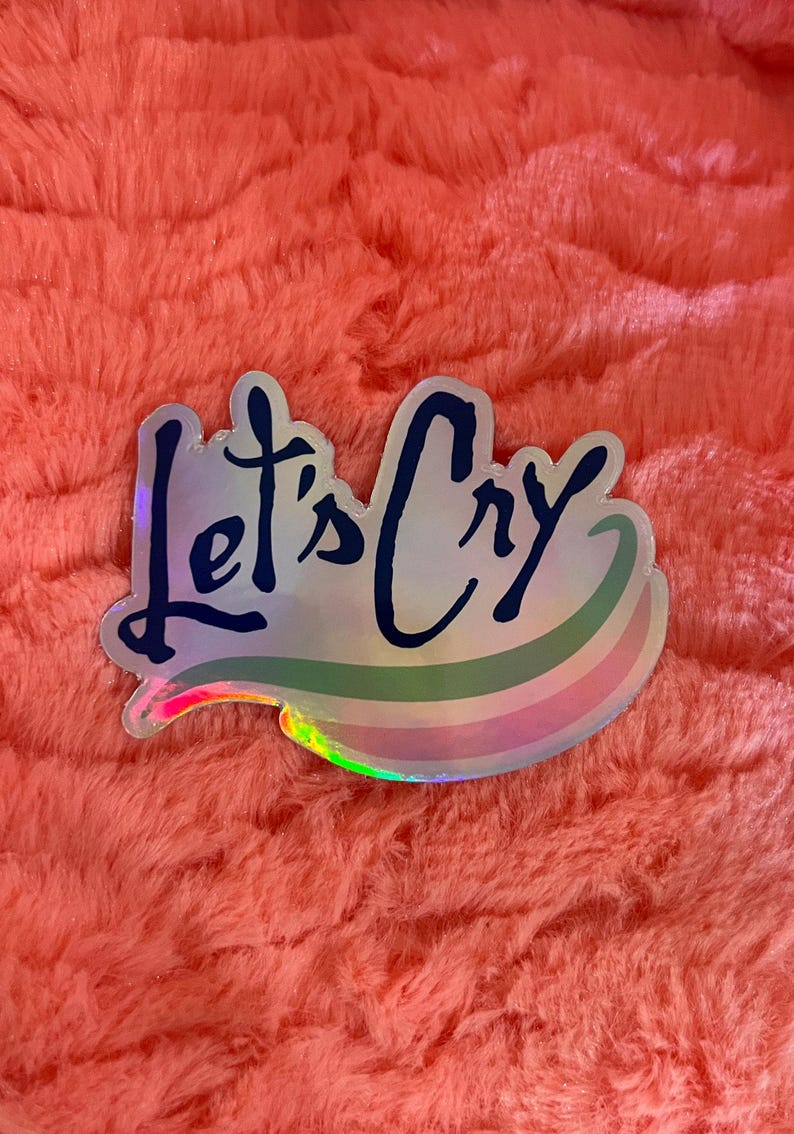 Peut inclure: Un autocollant holographique avec un motif arc-en-ciel et le texte "Let's Cry" en lettres noires.