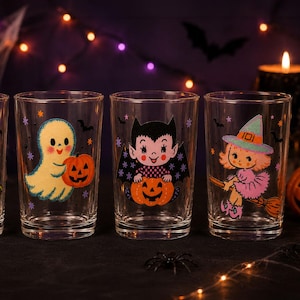 May include: Three clear glass tumblers featuring colorful Halloween-themed illustrations. The first glass shows a yellow ghost holding a pumpkin, the second a vampire with a pumpkin, and the third a witch on a broomstick with small stars and bats.