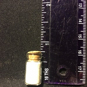May include: A small glass bottle with a cork stopper, filled with a white substance. The bottle is approximately 1.5 inches tall and is sitting next to a ruler.