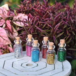 May include: Six small clear glass bottles with cork stoppers, each filled with a different colored glitter. The colors include silver, pink, blue, red, gold, and green. The bottles are arranged in a cluster on a light gray circular surface.