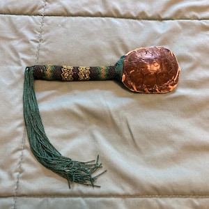 May include: A handcrafted turtle shell rattle with a beaded handle. The handle features a pattern of green, yellow, and black beads. A long strand of green beads hangs from the handle's end. The turtle shell is brown.