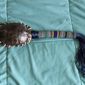 May include: A handcrafted ceremonial rattle featuring a turtle shell head, stitched with white thread. The handle is wrapped with blue and gray thread, adorned with a beaded pattern in green, blue, and orange, and finished with a blue leather fringe.