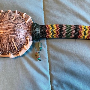 May include: A turtle shell rattle with a beaded handle. The handle is made of black, brown, green, yellow, and red beads. The handle has a long green tassel.