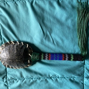 May include: A beaded turtle shell rattle with a green tassel. The handle is wrapped in green and black beads with blue, red, and white accents.