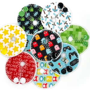 May include: A set of eight colorful fabric patches with various video game themes. The patches are round and have a hole in the center. The themes include Pac-Man ghosts, stars, Minecraft, game controllers, and pixelated spaceships.