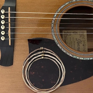 May include: Close-up of an acoustic guitar with a light brown wooden body and a dark brown pickguard. A set of new guitar strings is coiled on the pickguard. The guitar has a sound hole and a headstock with tuning pegs.