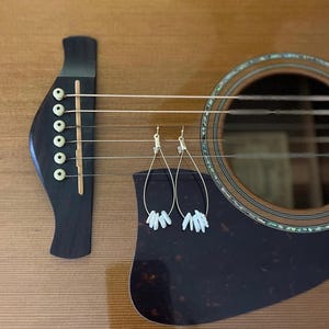 May include: Acoustic guitar with gold-colored teardrop earrings. The earrings have a cluster of white, irregular-shaped beads. The guitar is brown with a dark pickguard and strings.