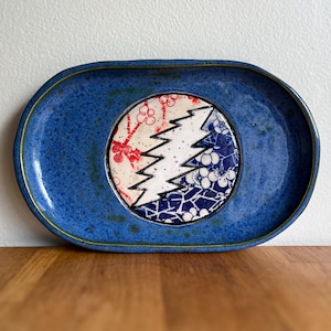 May include: A blue ceramic serving tray with a central design featuring a white lightning bolt and floral patterns in red and blue. The tray has an oval shape and a textured surface, suitable for serving food or displaying decorative items.