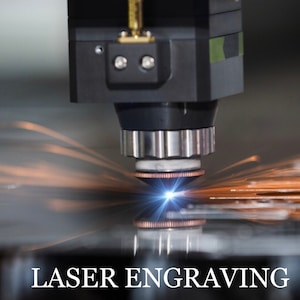 May include: Close-up of a laser engraving machine in operation. A bright blue laser beam is focused on a metal surface, creating sparks and smoke. The text "LASER ENGRAVING" is visible at the bottom of the image.