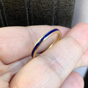 Puede incluir: Un anillo de oro con una fina banda de esmalte azul vibrante. El anillo se sostiene en una mano, mostrando su dise&ntilde;o simple pero elegante. La superficie pulida del anillo refleja la luz, resaltando el contraste entre el oro y el azul.