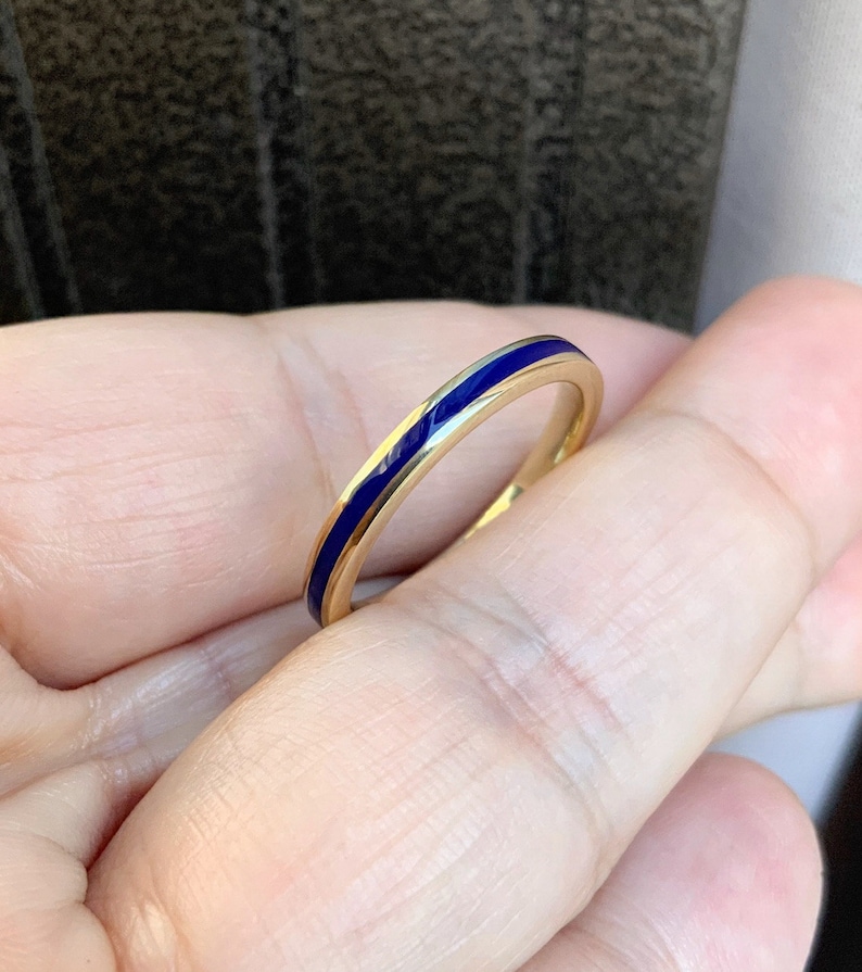 Puede incluir: Un anillo de oro con una fina banda de esmalte azul vibrante. El anillo se sostiene en una mano, mostrando su dise&ntilde;o simple pero elegante. La superficie pulida del anillo refleja la luz, resaltando el contraste entre el oro y el azul.