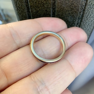 Puede incluir: Un anillo de banda de oro con un dise&ntilde;o delgado y pulido. El anillo tiene una superficie lisa y reflectante y una banda interior oscura, posiblemente con incrustaciones. El anillo se sostiene en una mano, mostrando su simple elegancia.