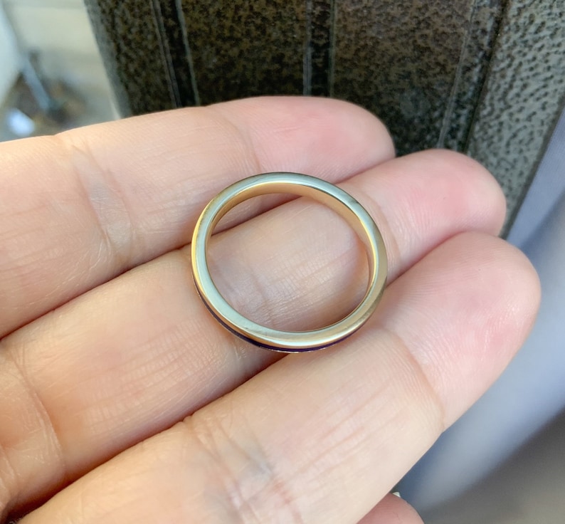 Puede incluir: Un anillo de banda de oro con un dise&ntilde;o delgado y pulido. El anillo tiene una superficie lisa y reflectante y una banda interior oscura, posiblemente con incrustaciones. El anillo se sostiene en una mano, mostrando su simple elegancia.