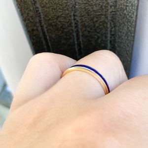 Puede incluir: Un anillo de oro con una banda delgada y una llamativa franja de esmalte azul. El anillo es simple y elegante, perfecto para el uso diario. El anillo se muestra en un dedo.