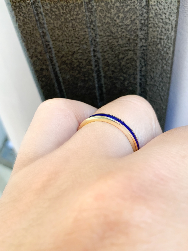 Puede incluir: Un anillo de oro con una banda delgada y una llamativa franja de esmalte azul. El anillo es simple y elegante, perfecto para el uso diario. El anillo se muestra en un dedo.