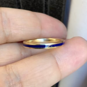 Puede incluir: Un anillo de banda dorado con una fina y vibrante franja de esmalte azul. El anillo se sostiene entre los dedos, mostrando su acabado pulido y su dise&ntilde;o simple y elegante. Esta joya es adecuada para el uso diario.