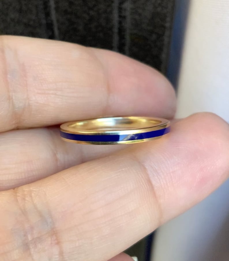 Puede incluir: Un anillo de banda dorado con una fina y vibrante franja de esmalte azul. El anillo se sostiene entre los dedos, mostrando su acabado pulido y su dise&ntilde;o simple y elegante. Esta joya es adecuada para el uso diario.