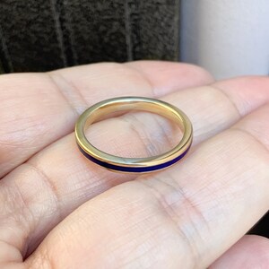 Puede incluir: Un anillo de oro con una fina banda de esmalte azul oscuro. El anillo es sencillo y elegante, con un acabado pulido. El anillo se muestra en una mano.