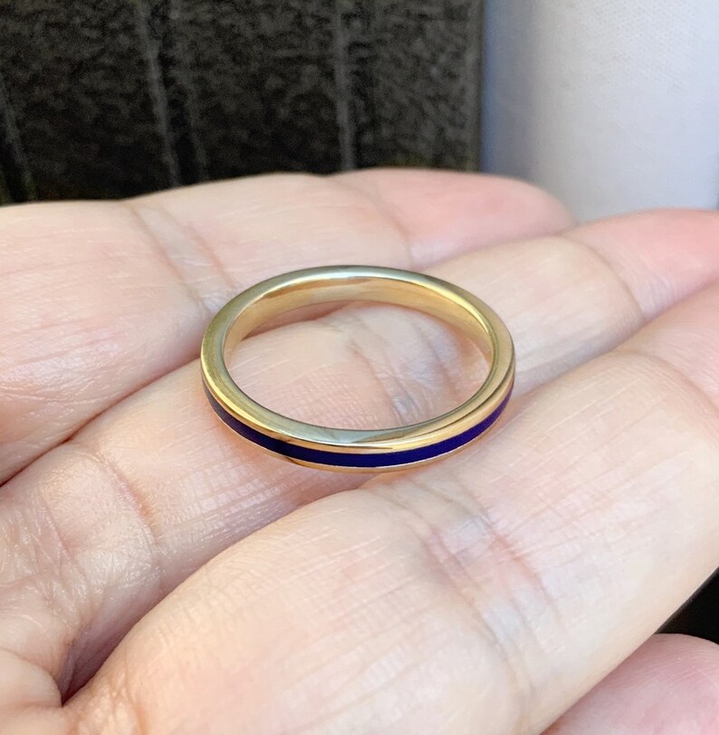 Puede incluir: Un anillo de oro con una fina banda de esmalte azul oscuro. El anillo es sencillo y elegante, con un acabado pulido. El anillo se muestra en una mano.