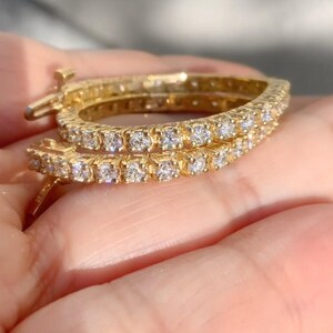 May include: A pair of gold hoop earrings with a row of small, clear cubic zirconia stones around the edge of each hoop.