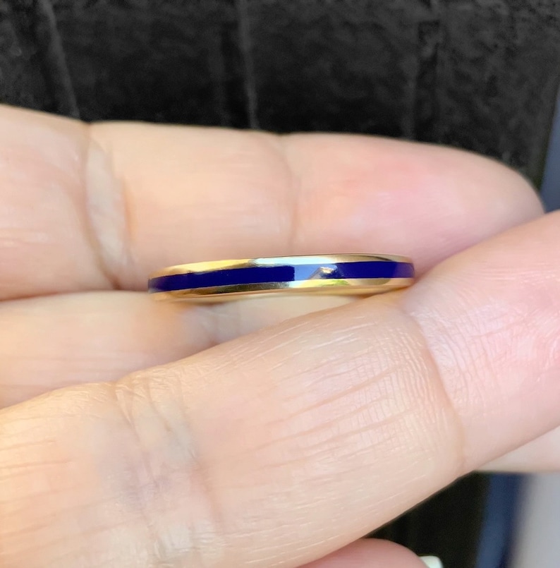 Puede incluir: Un anillo de oro con una fina banda de esmalte azul brillante. El anillo se sostiene en una mano, centr&aacute;ndose en el dise&ntilde;o y el color del anillo.