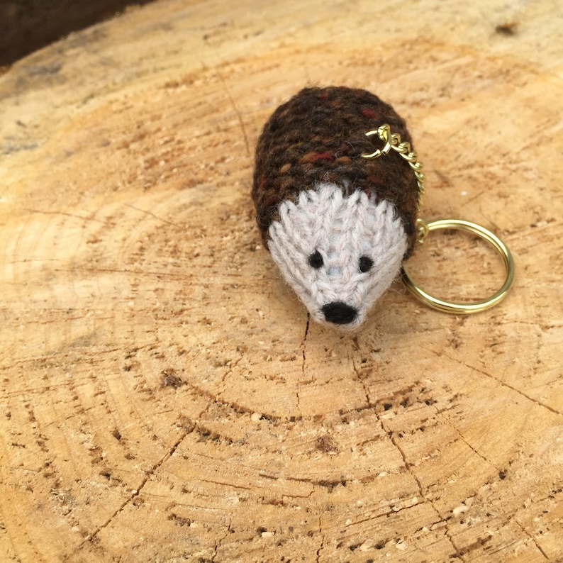 K&ouml;nnte beinhalten: Ein brauner und wei&szlig;er gestrickter Igel-Schl&uuml;sselanh&auml;nger mit einem goldenen Schl&uuml;sselring. Der Igel sitzt auf einer Holzoberfl&auml;che.
