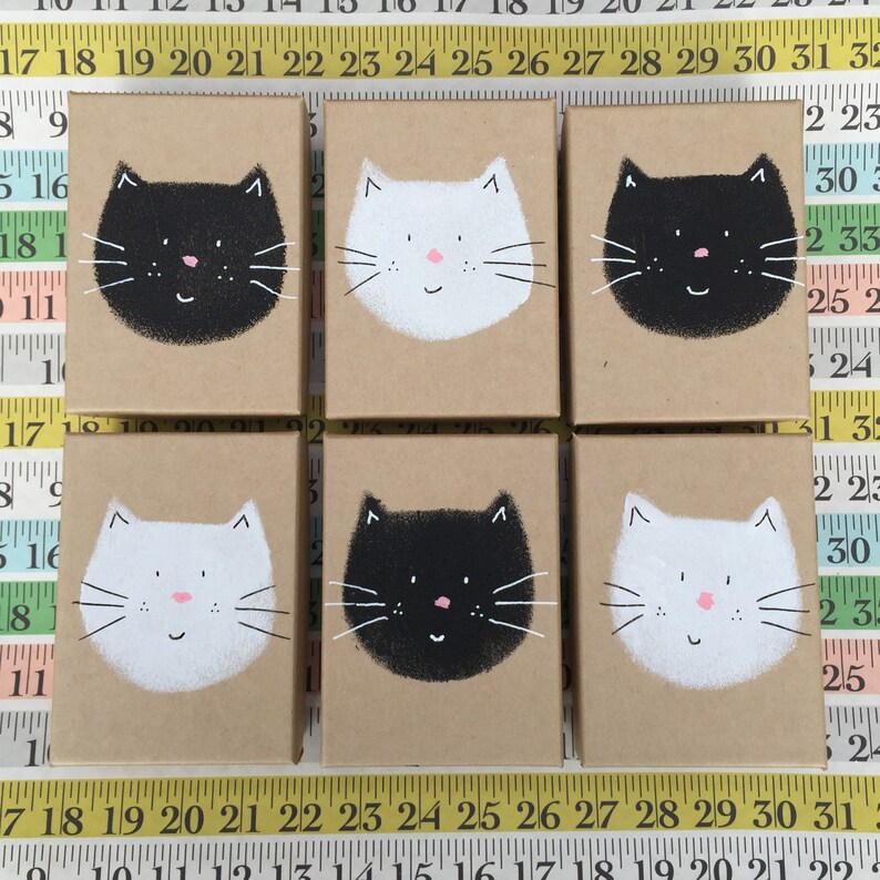 Puede incluir: Seis peque&ntilde;as cajas de cart&oacute;n con caras de gato pintadas. Tres cajas tienen gatos negros con caras blancas y tres cajas tienen gatos blancos con caras negras.