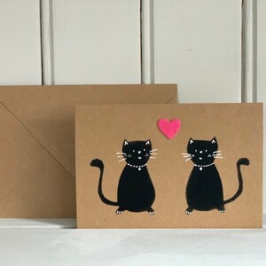 May include: A greeting card with two black cat illustrations, each wearing a white collar, and a pink heart above them. The card is on a brown background with a matching envelope.