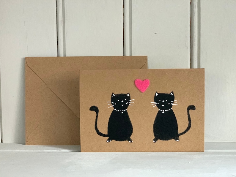 May include: A greeting card with two black cat illustrations, each wearing a white collar, and a pink heart above them. The card is on a brown background with a matching envelope.