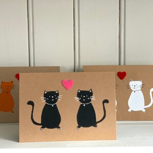May include: Three handmade greeting cards with cat illustrations. One card features an orange cat, another has two black cats, and the third shows a white cat. Each card has a felt heart accent. The cards are on a white shelf.