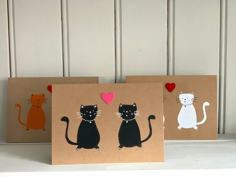 May include: Three handmade greeting cards with cat illustrations. One card features an orange cat, another has two black cats, and the third shows a white cat. Each card has a felt heart accent. The cards are on a white shelf.