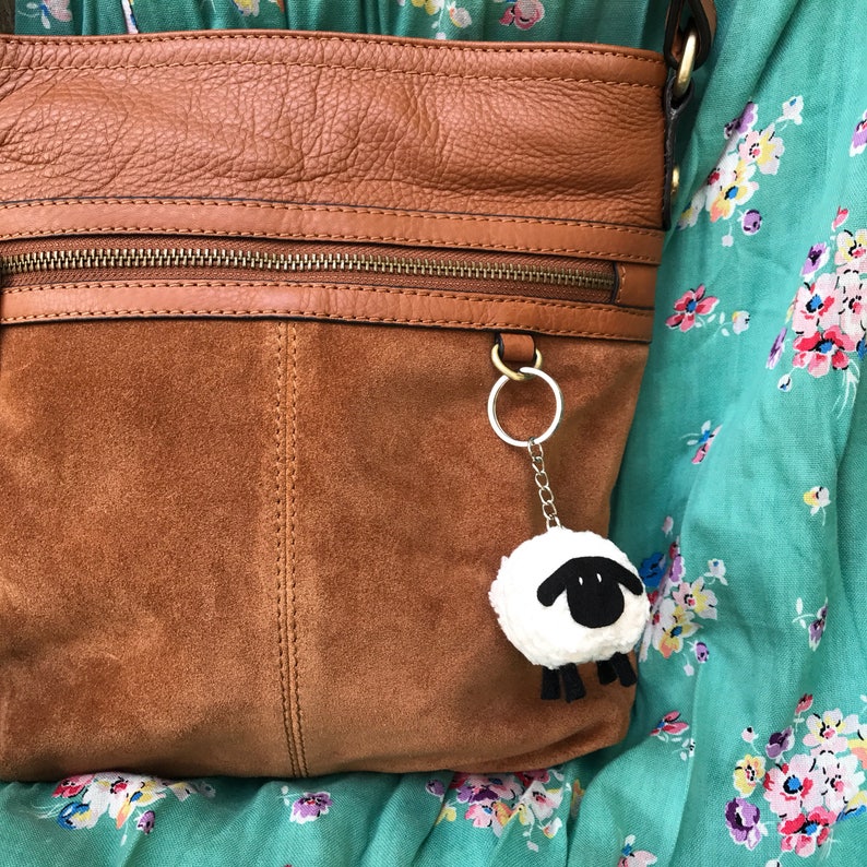 May include: A brown leather and suede handbag with a zipper closure and a fluffy white sheep keychain.