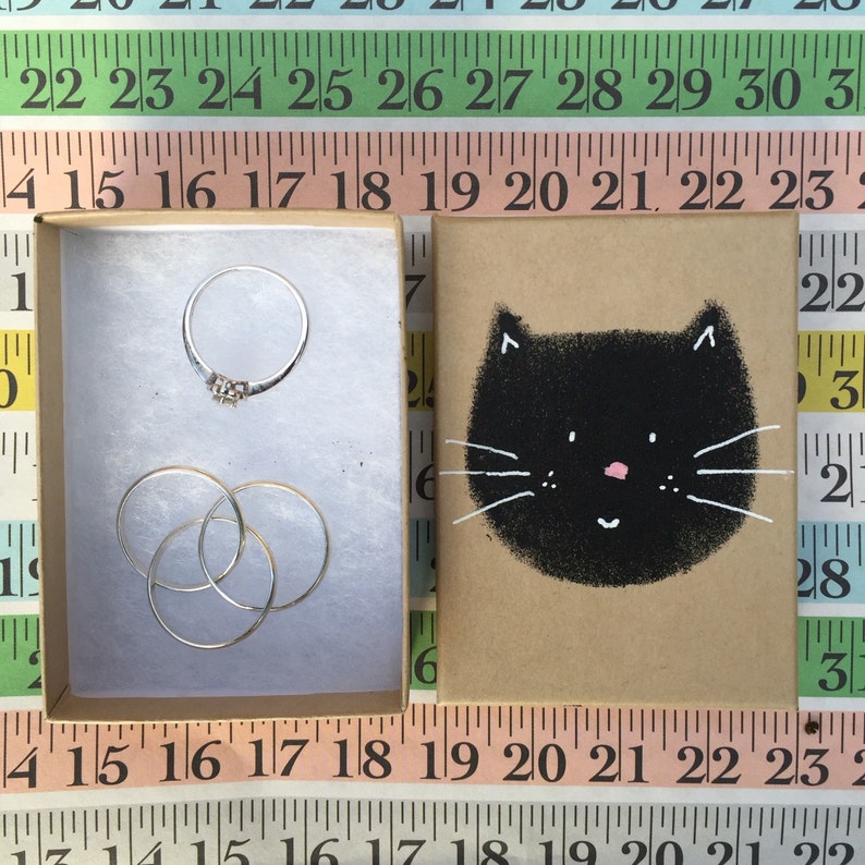 Puede incluir: Dos cajas peque&ntilde;as, una con un anillo de plata y tres anillos de plata, la otra con una ilustraci&oacute;n de un gato negro sobre un fondo marr&oacute;n.