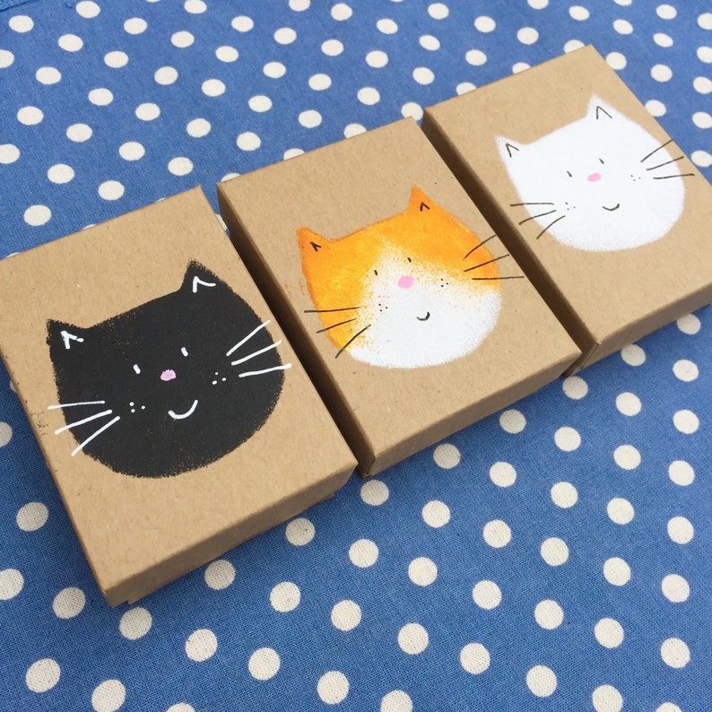 Puede incluir: Tres peque&ntilde;as cajas de cart&oacute;n con caras de gato pintadas. Las cajas son marrones y tienen una cara de gato blanca con bigotes negros, una cara de gato naranja con bigotes blancos y una cara de gato negra con bigotes blancos.
