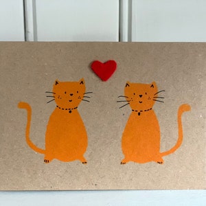 May include: A greeting card with a kraft paper background features two orange cartoon cats with black whiskers and collars. A small red heart is above the cats. The card is a simple, cute design.