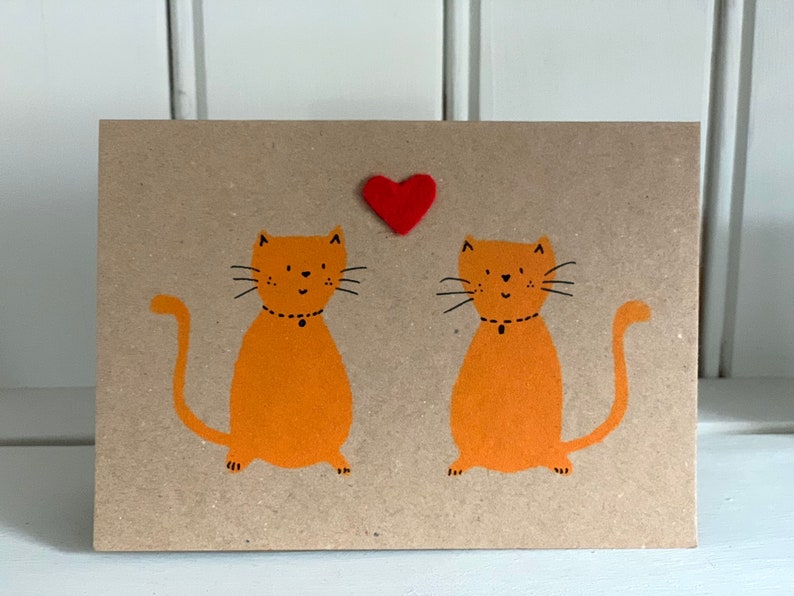 May include: A greeting card with a kraft paper background features two orange cartoon cats with black whiskers and collars. A small red heart is above the cats. The card is a simple, cute design.
