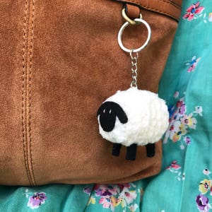 May include: A white crocheted sheep keychain with black eyes and legs attached to a silver key ring. The keychain is hanging from a brown leather bag.