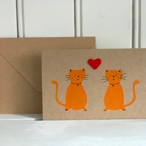 May include: A greeting card with two orange cartoon cats and a red heart. The cats have black whiskers and are wearing black collars. A brown envelope is next to the card. The card and envelope are on a white surface.