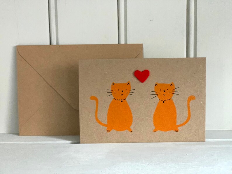 May include: A greeting card with two orange cartoon cats and a red heart. The cats have black whiskers and are wearing black collars. A brown envelope is next to the card. The card and envelope are on a white surface.