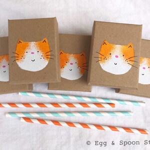 Puede incluir: Cinco peque&ntilde;as cajas marrones con caras de gato pintadas de naranja y blanco en la parte delantera. Las cajas est&aacute;n dispuestas en fila sobre una superficie blanca. Hay cinco pajitas rayadas de naranja y blanco junto a las cajas.