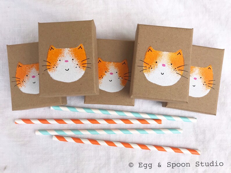 Puede incluir: Cinco peque&ntilde;as cajas marrones con caras de gato pintadas de naranja y blanco en la parte delantera. Las cajas est&aacute;n dispuestas en fila sobre una superficie blanca. Hay cinco pajitas rayadas de naranja y blanco junto a las cajas.